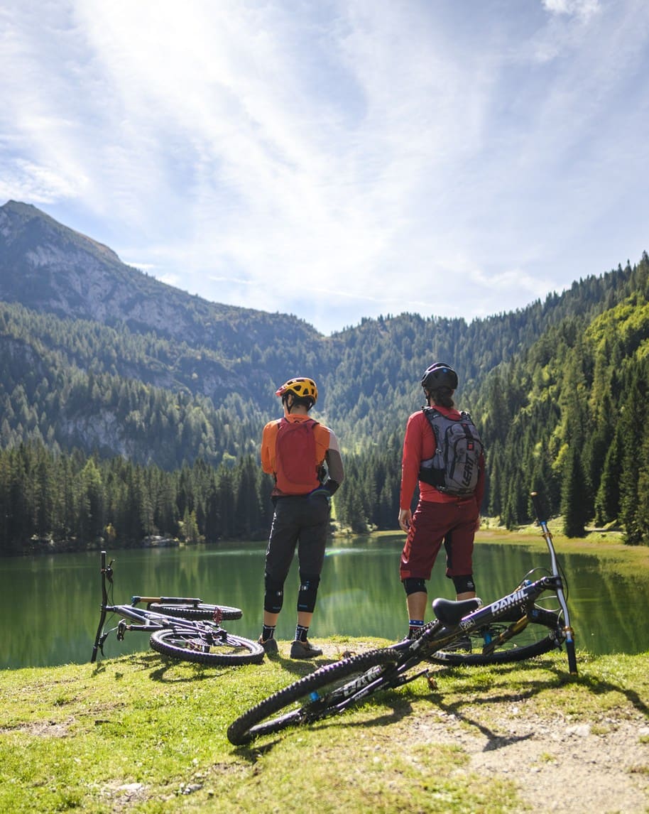 Due ciclisti con casco e zaino in piedi sulla riva di un lago montano, biciclette sdraiate sul prato