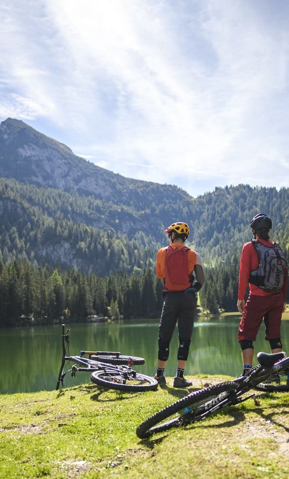 Due ciclisti in abbigliamento tecnico in piedi sulla riva di un lago alpino, biciclette appoggiate sull'erba
