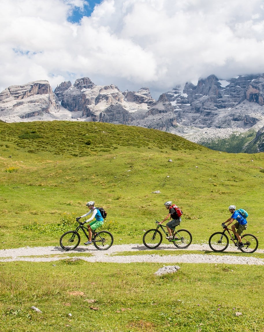 Tre ciclisti in fila pedalano su un sentiero ghiaioso attraverso un prato alpino con montagne rocciose sullo sfondo