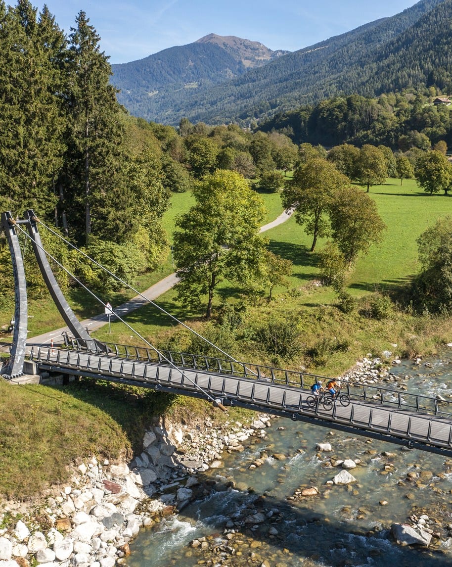Ponte pedonale sospeso con due ciclisti che attraversano un fiume, prati verdi e montagne sullo sfondo