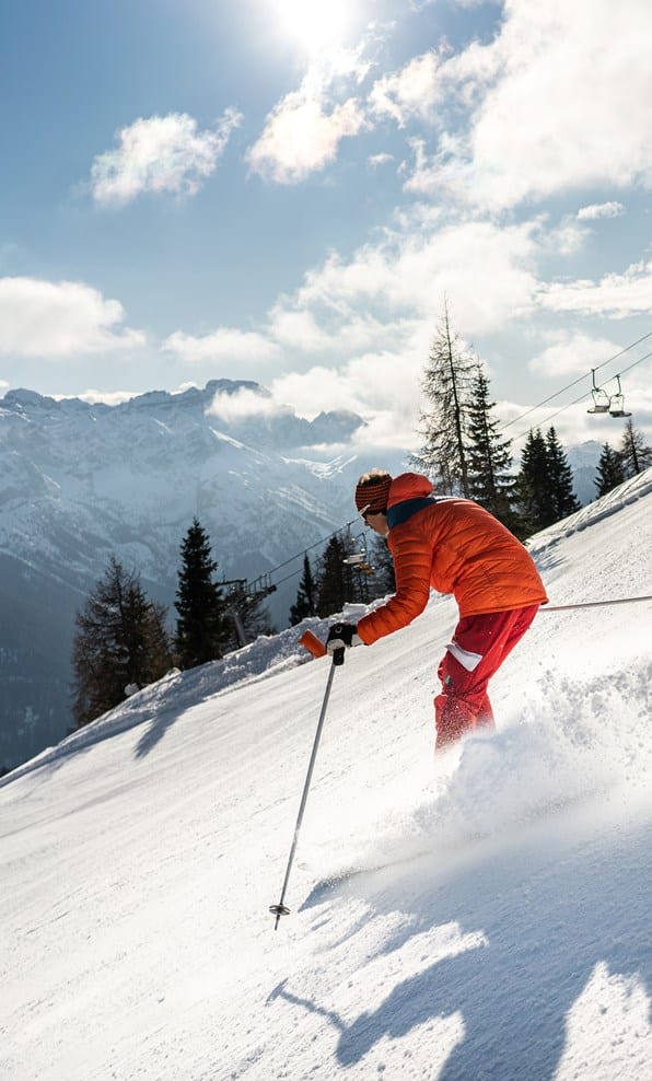 Sciatore con giacca arancione e pantaloni rossi scende su pista innevata con montagne e seggiovia sullo sfondo