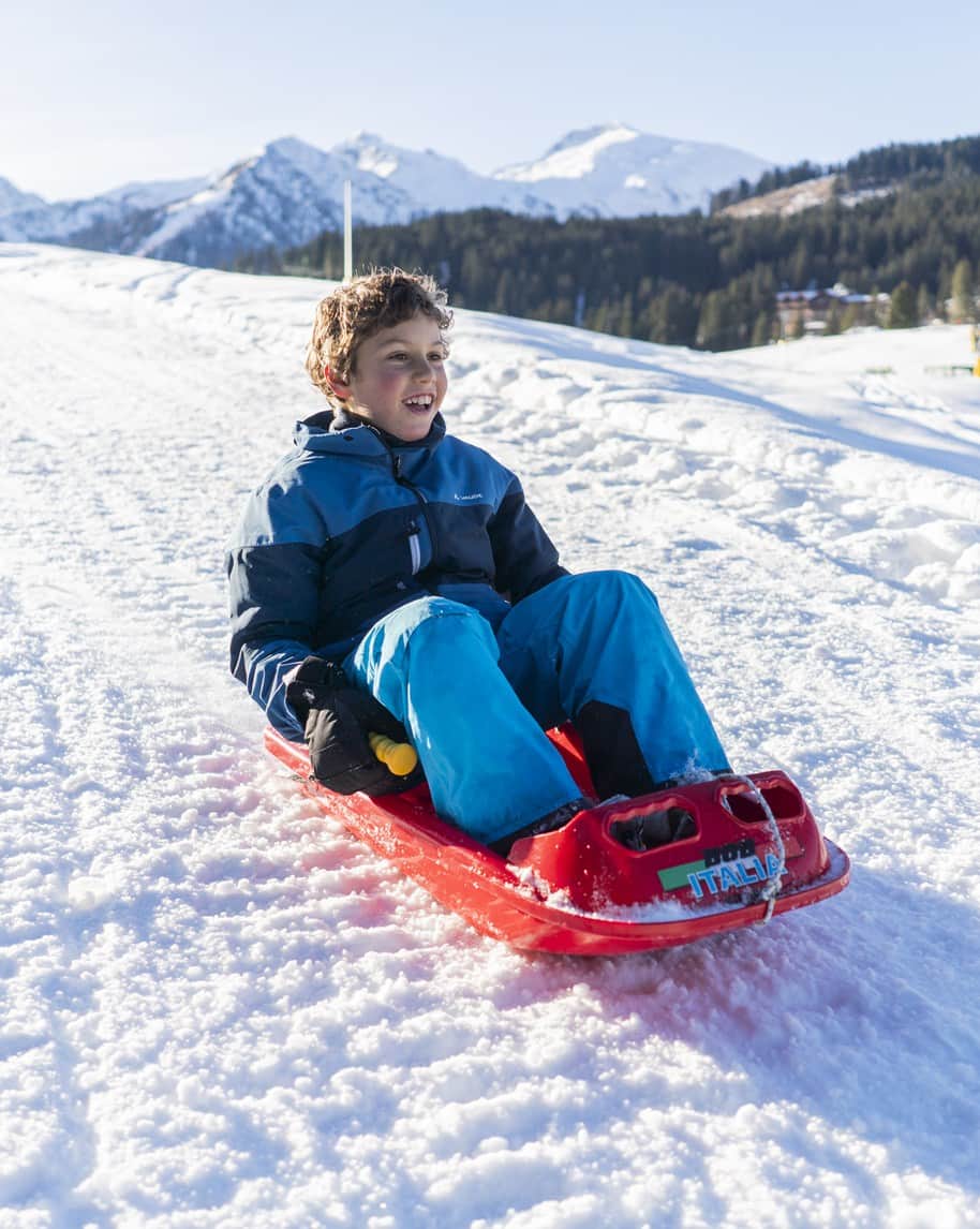 Bambino sorridente seduto su slitta rossa che scende su neve con montagne innevate sullo sfondo