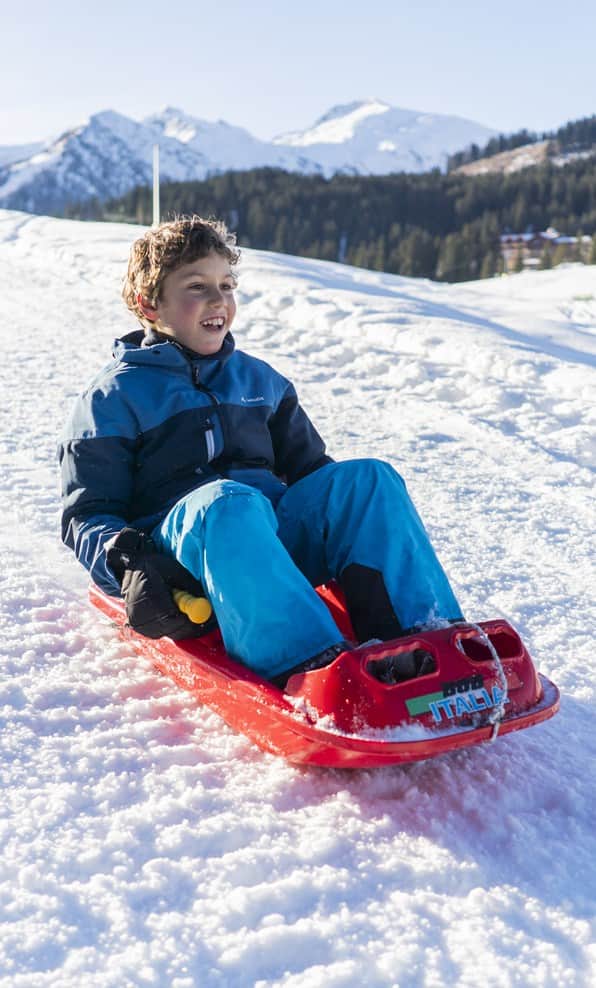 Bambino seduto su una slitta rossa che scivola sulla neve, sorridente con montagne innevate sullo sfondo