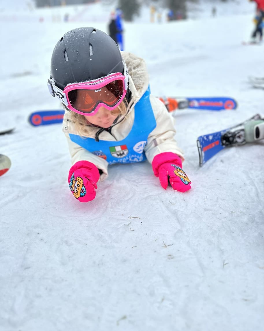 Bambino con casco e maschera rosa sdraiato sulla neve, guanti rosa e sci ai lati