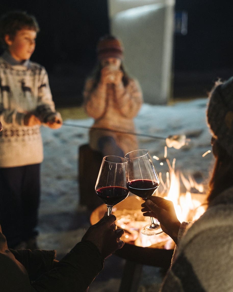 Due persone brindano con calici di vino rosso davanti a un falò sulla neve; bambini sullo sfondo