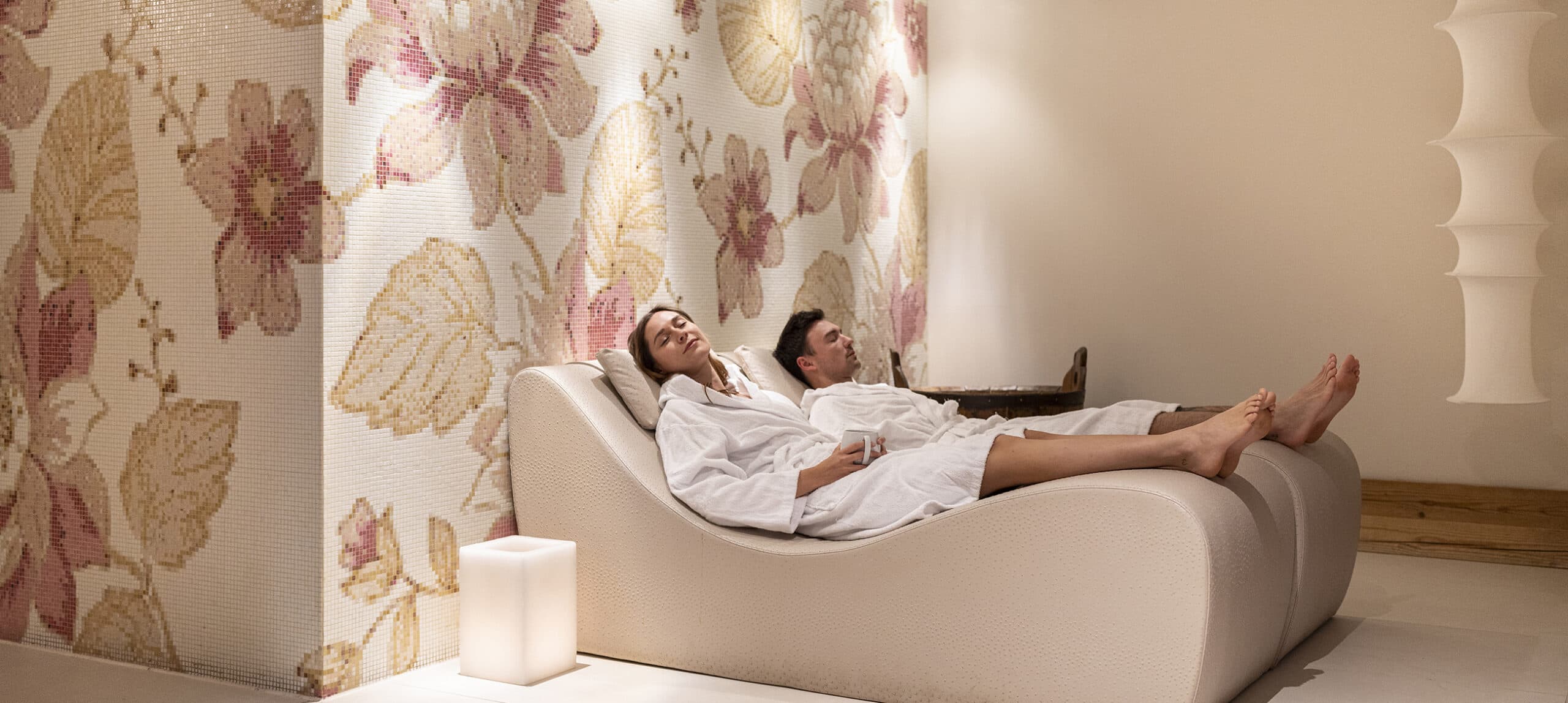 Coppia in accappatoi sdraiata su chaise longue in spa con parete mosaico floreale e luce soffusa