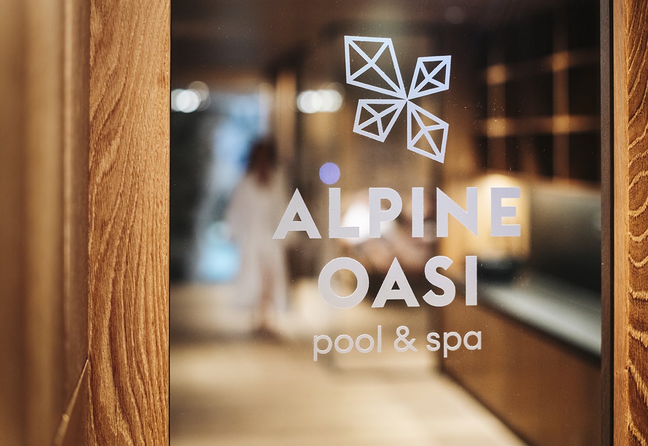 Vetro con scritta ALPINE OASI pool & spa, dietro corridoio spa sfocato e figura in accappatoio