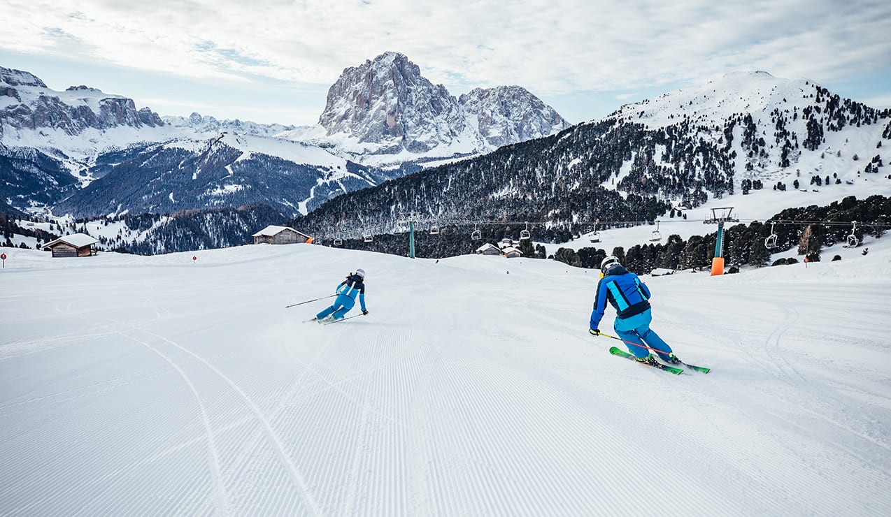 Due sciatori in tuta blu scendono una pista battuta, seggiovia e montagne innevate sullo sfondo