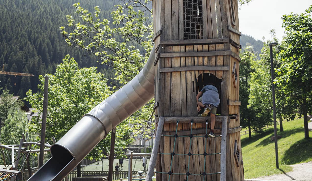 Torre gioco in legno con scivolo metallico; bambino arrampica entrando, alberi e montagne sullo sfondo