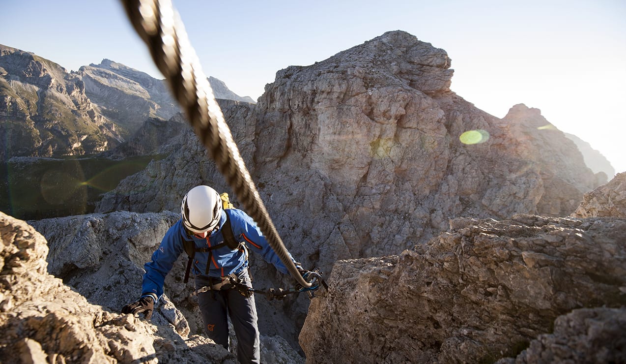 Alpinista con casco e giacca blu risale via ferrata aggrappato a un cavo su parete rocciosa