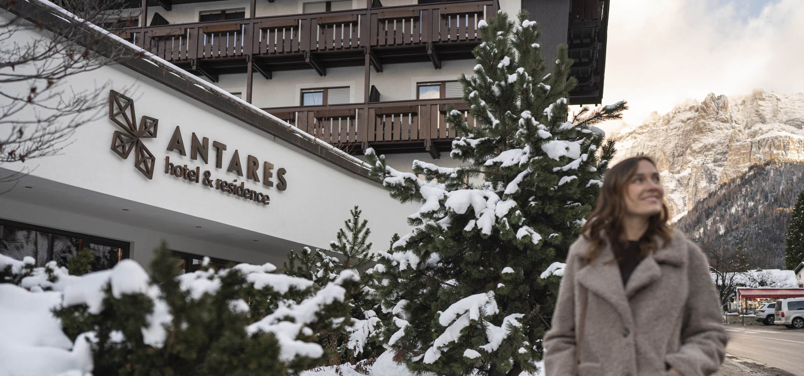 Antares hotel & residence su edificio innevato; donna in cappotto in primo piano e montagne sullo sfondo