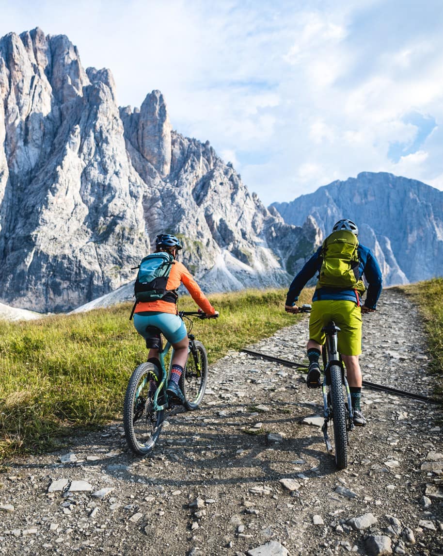 Due ciclisti con casco e zaini salgono in mountain bike su sentiero roccioso verso alte pareti montuose
