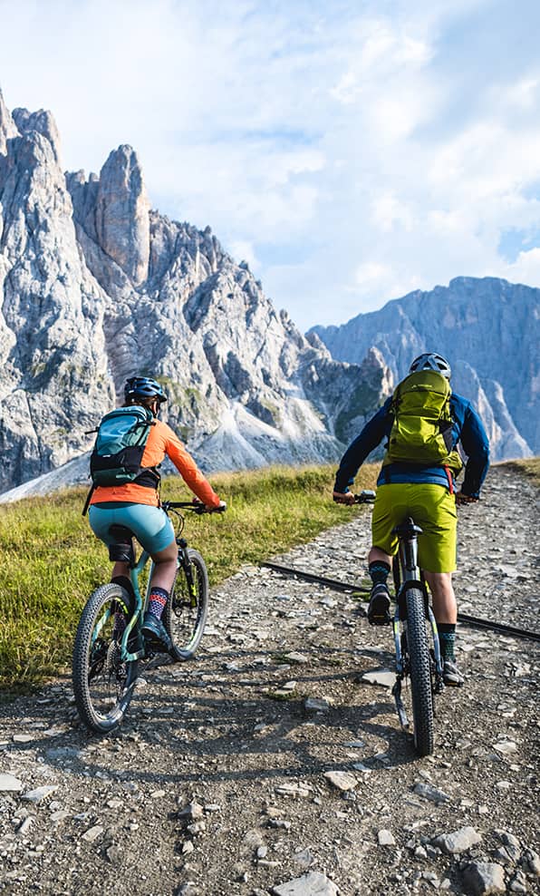 Due ciclisti con zaini salgono su sentiero di montagna verso ripide pareti rocciose