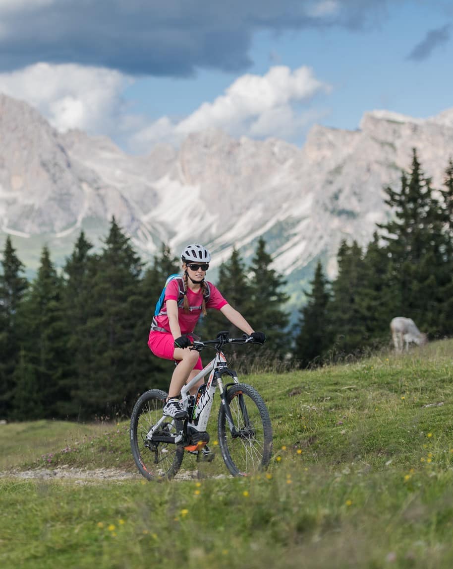 Ciclista in abbigliamento rosa e casco su mountain bike su sentiero alpino, pineta e montagne rocciose sullo sfondo