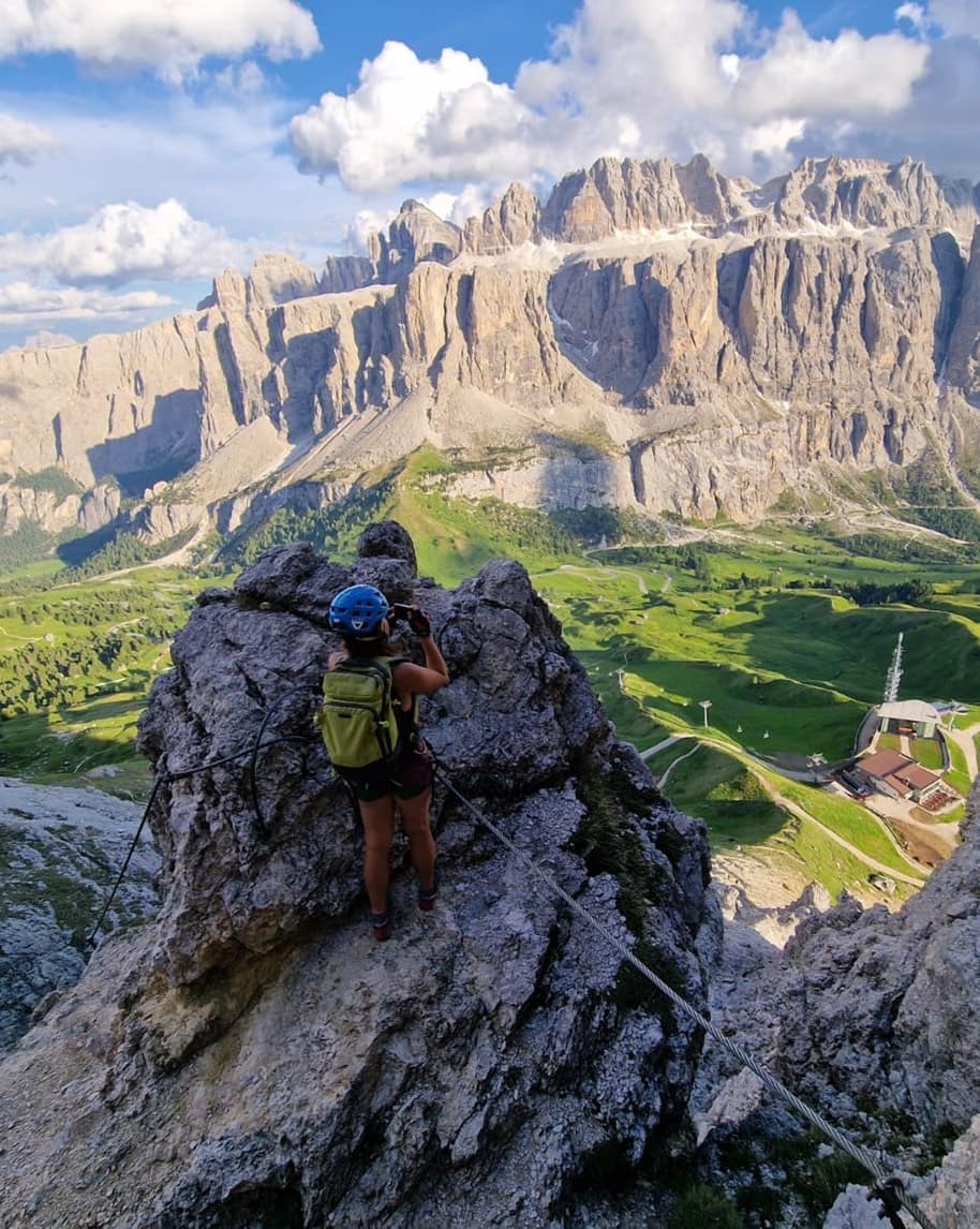 Persona con casco e zaino su via ferrata agganciata al cavo fotografa la valle verde e le pareti rocciose