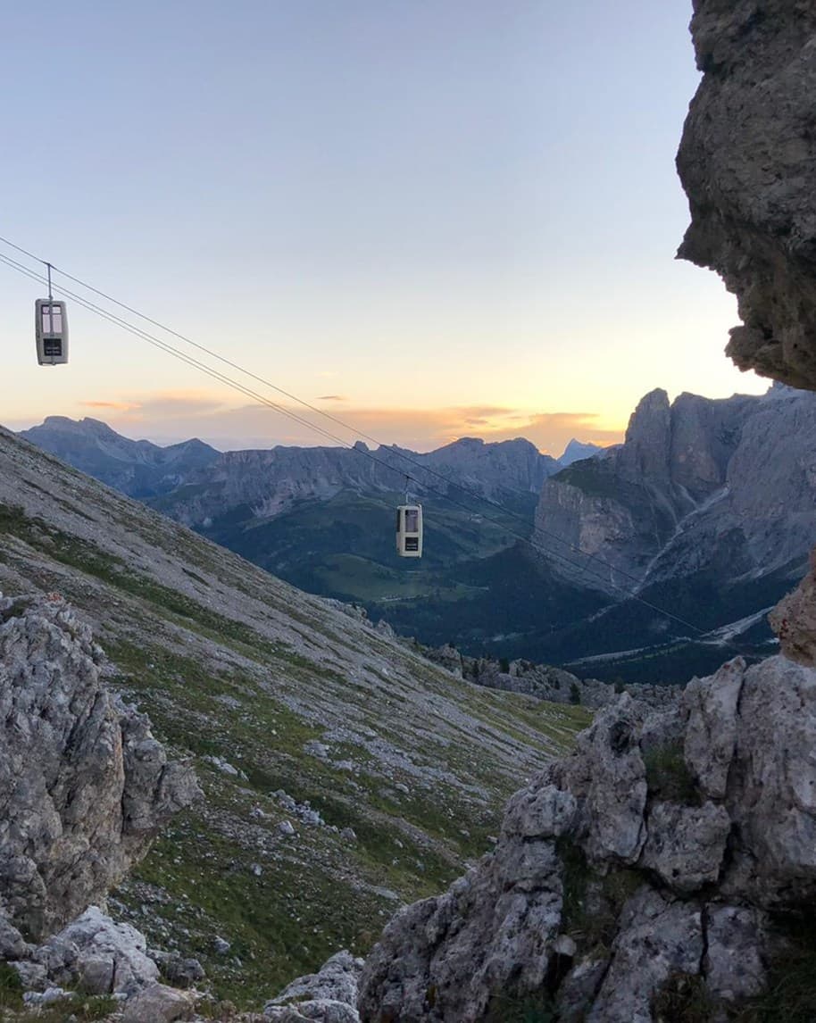 Due cabine di funivia sospese su cavi sopra un pendio roccioso, vallata e cime montane al tramonto