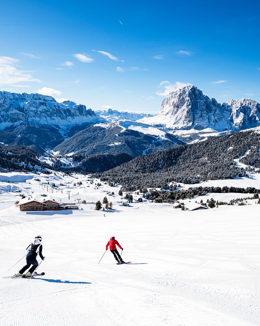 Due sciatori in discesa su pista innevata, baite in basso e massiccio montuoso sullo sfondo