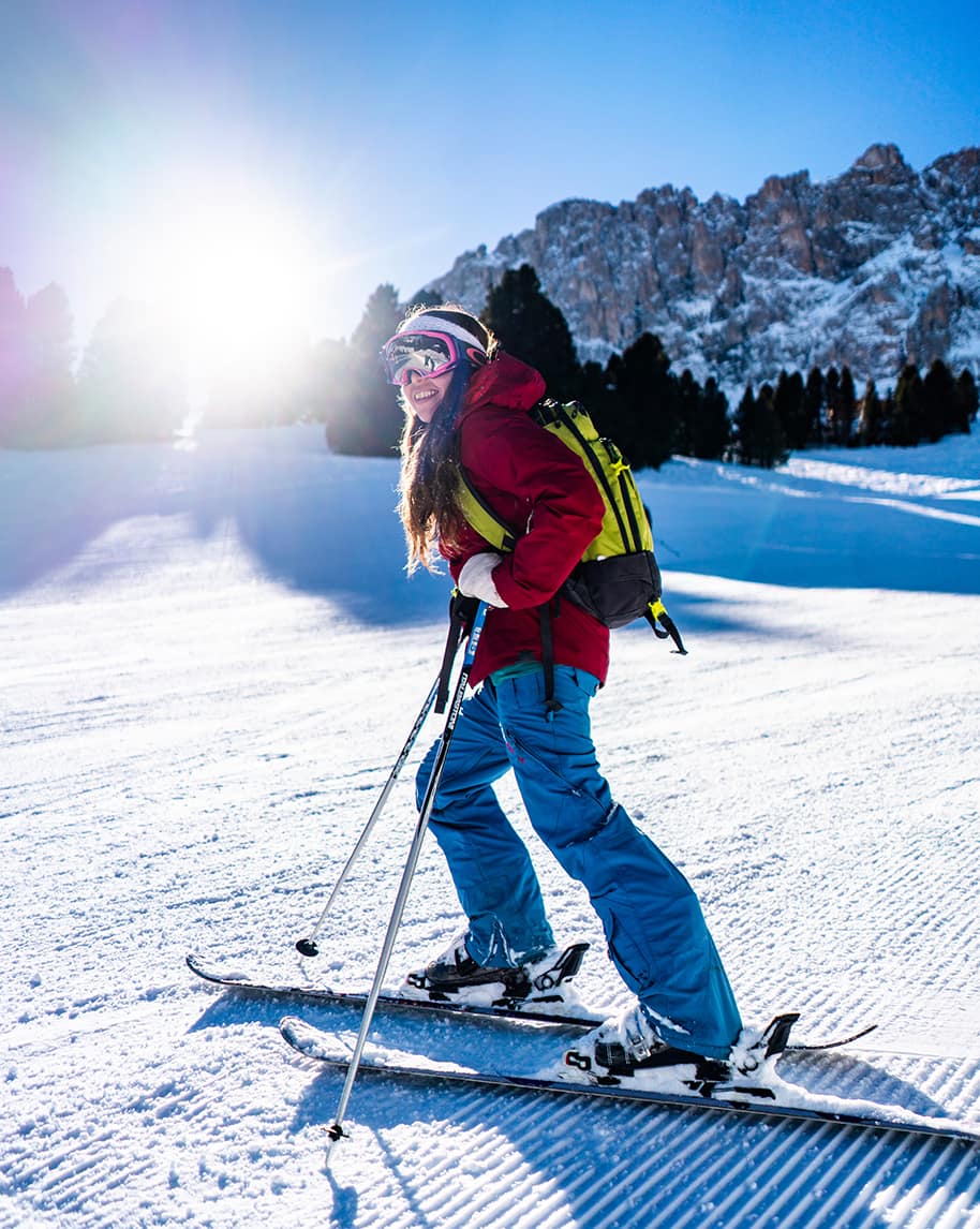 Sciatrice con giacca rossa e zaino giallo scia su pista battuta, sole basso e montagne innevate sullo sfondo