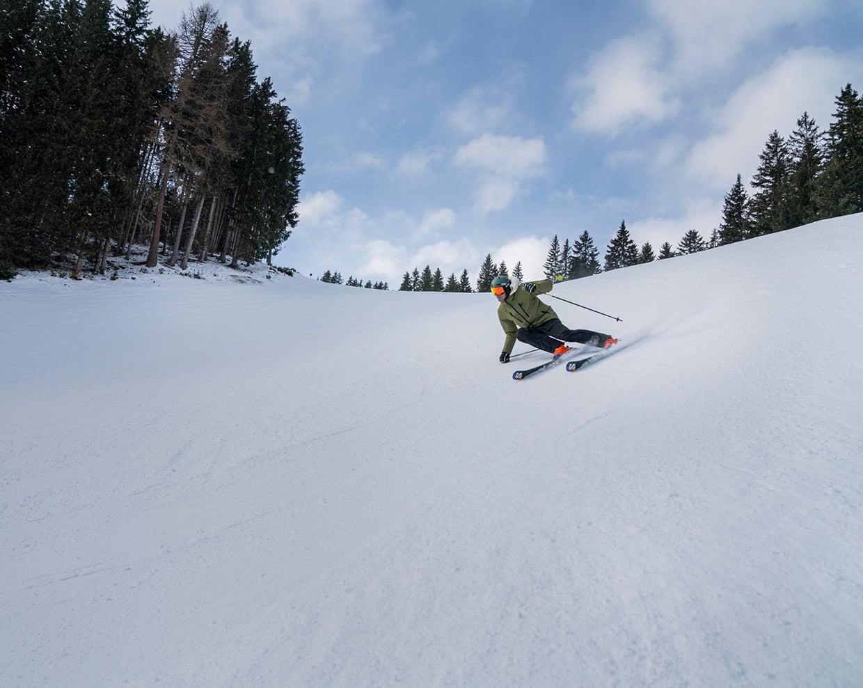 Sciatore in giacca verde curva su pista innevata, alberi di conifere sullo sfondo