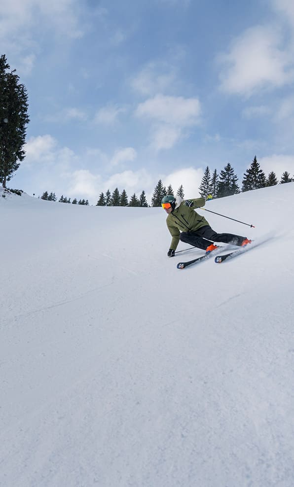 Sciatore con giacca verde curva sul pendio innevato, scia di neve, alberi e cielo sullo sfondo