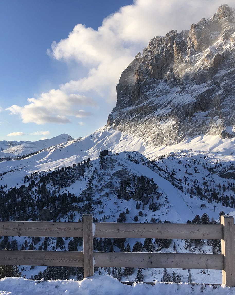 Recinzione in legno in primo piano, piccola baita su cresta innevata e alta parete rocciosa sotto cielo azzurro