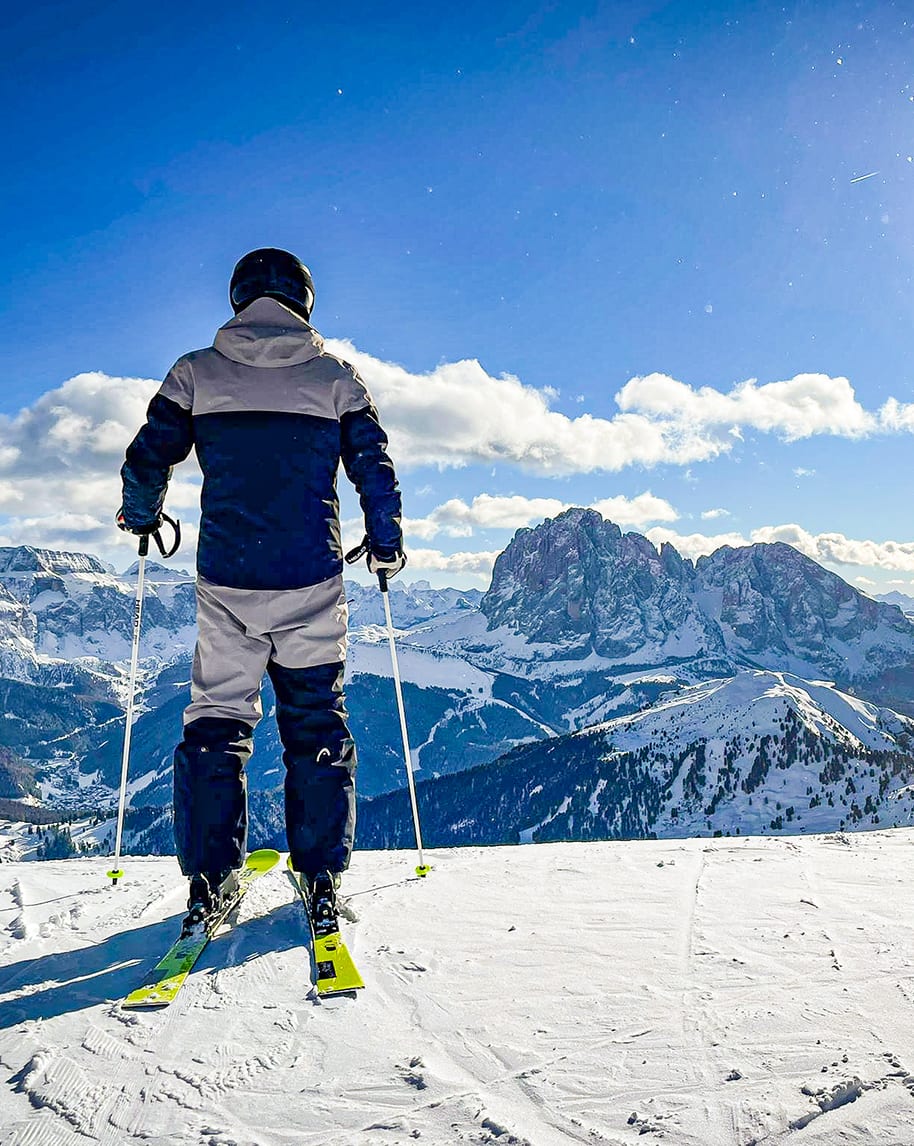 Sciatore di spalle in tuta e casco, fermo con sci e bastoncini su pendio innevato, vista su montagne