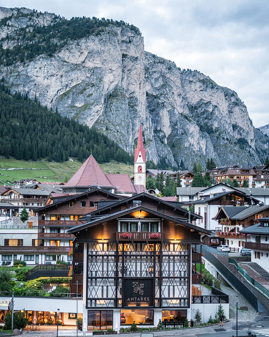 Edificio Hotel Antares in paese alpino, davanti a chiesa con alto campanile e parete rocciosa.