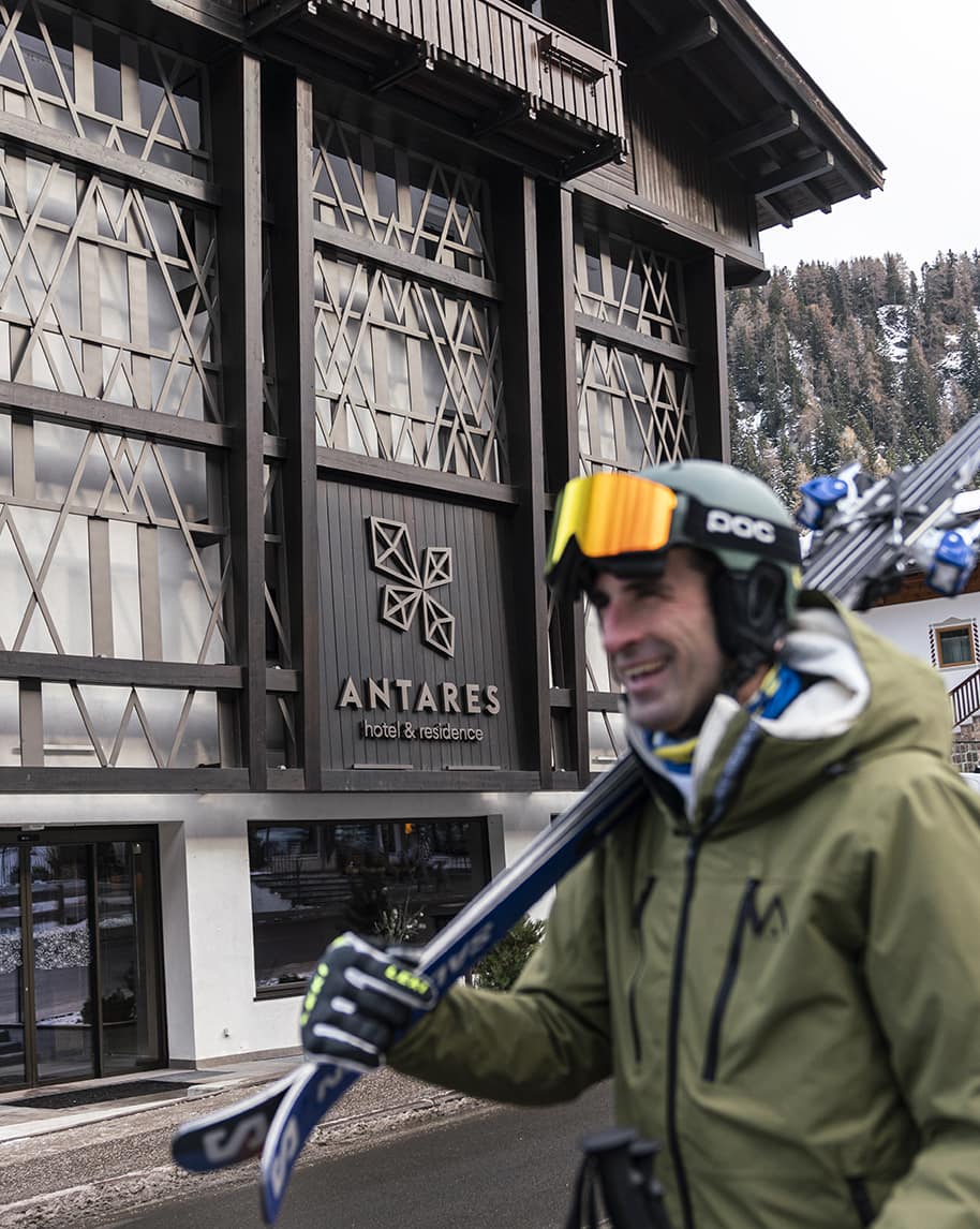 Antares hotel & residence sullo sfondo, sciatore con casco e sci in spalla in primo piano