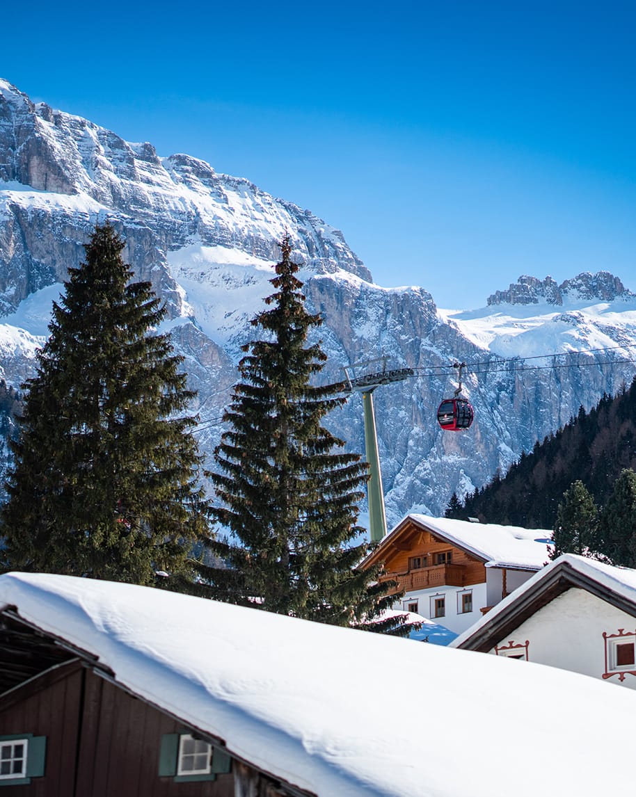 Cabina della funivia rossa sospesa sopra case e abeti innevati, con alte montagne rocciose e cielo blu sullo sfondo