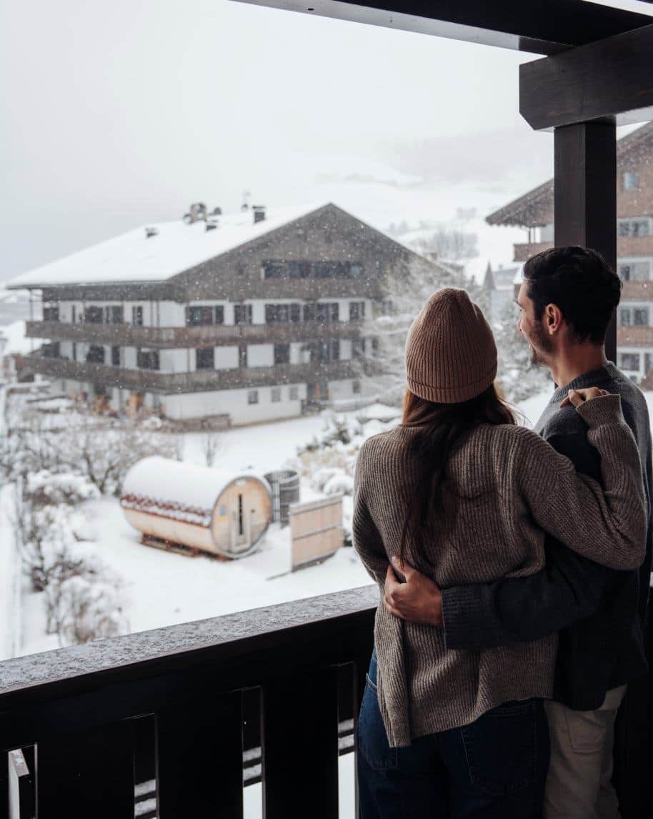 Coppia abbracciata su balcone che guarda paesaggio innevato con chalet alpini