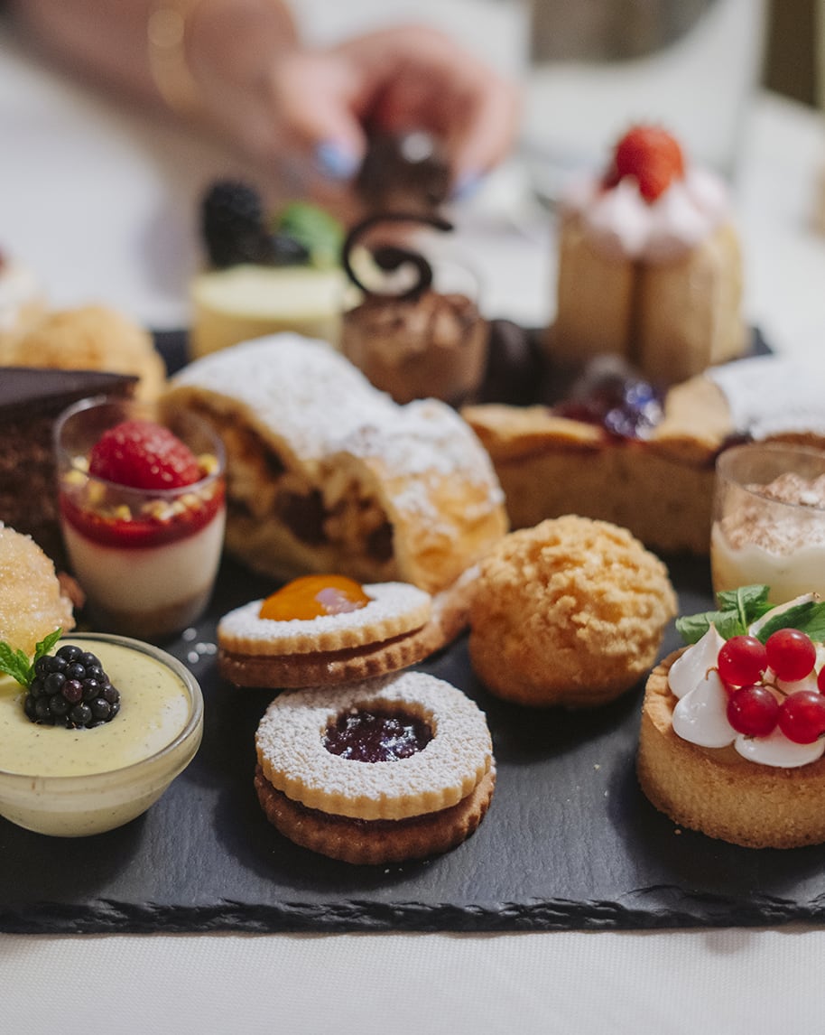 Vassoio scuro con pasticcini assortiti (biscotti con marmellata, bigné, tartellette), mano sfocata sullo sfondo