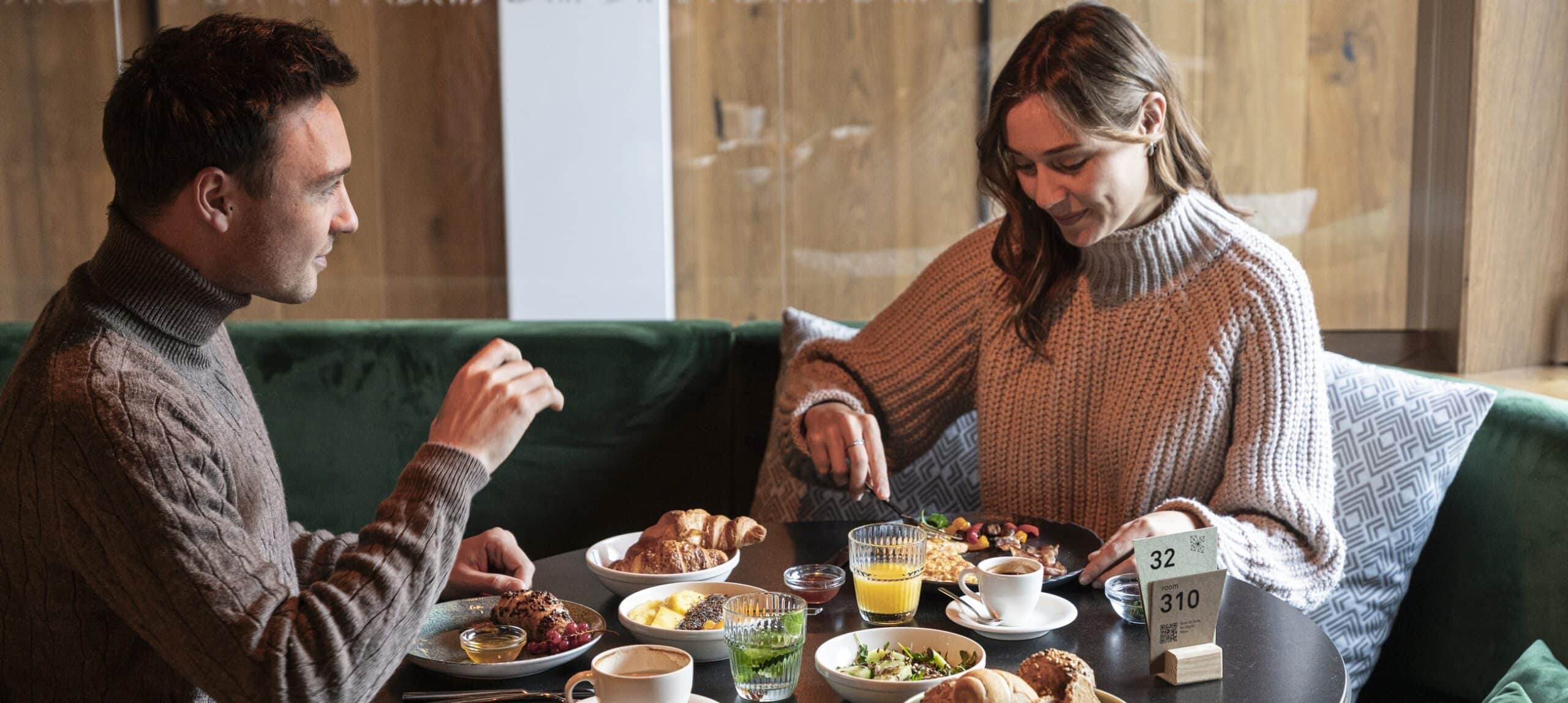 Coppia seduta a un tavolo che fa colazione in un locale, piatti e cartello con scritto '32 room 310'