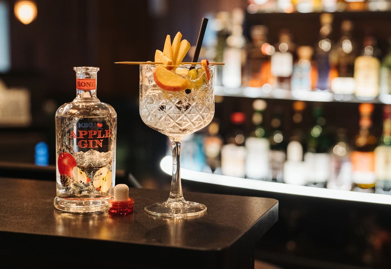 Bicchiere da cocktail con fettine di mela e cannuccia accanto a bottiglia APPLE GIN su bancone di bar