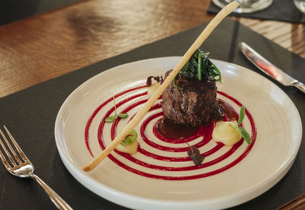Medaglione di carne con spinaci su salsa rossa a spirale, purea e grissino su piatto bianco