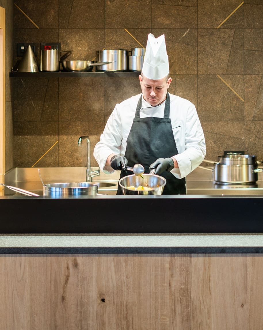Cuoco con cappello da chef e grembiule versa cibo con un mestolo in cucina professionale