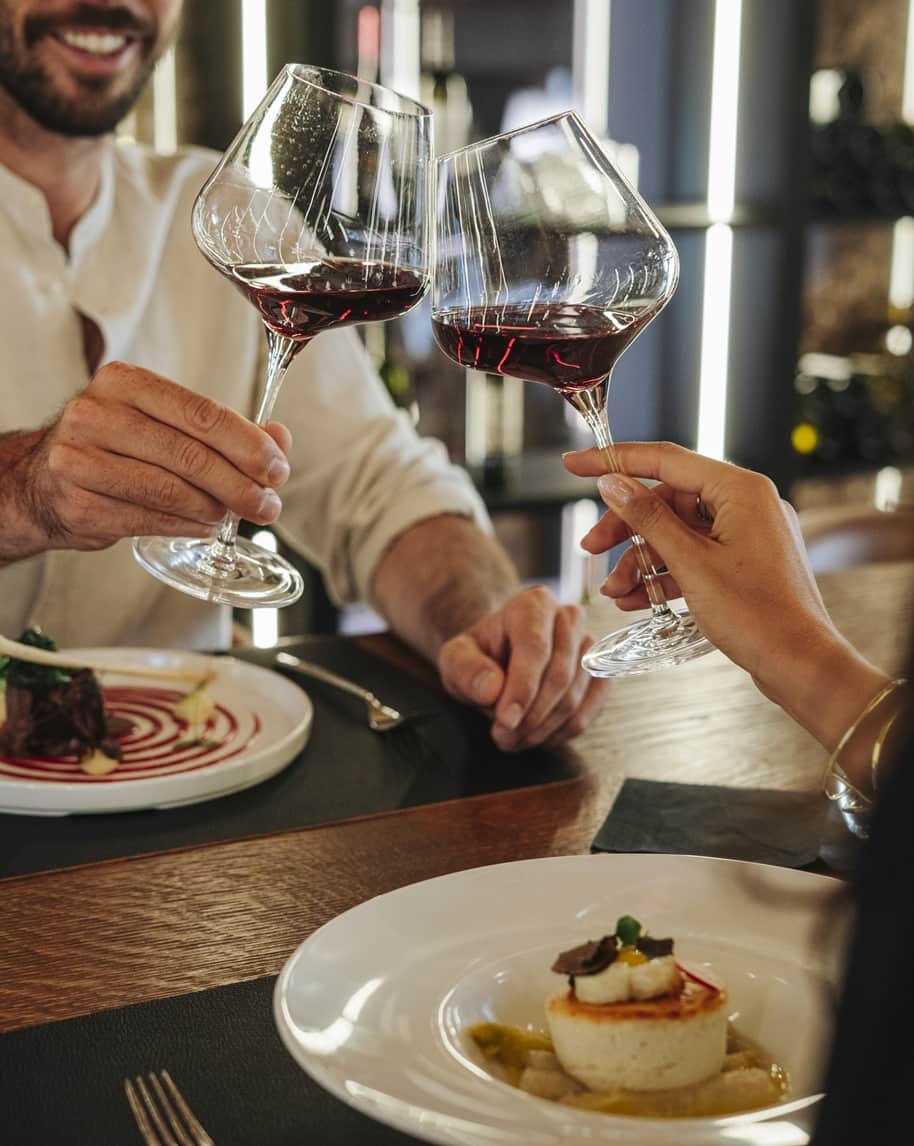 Due calici di vino rosso che brindano sopra un tavolo con piatti e mani che li reggono