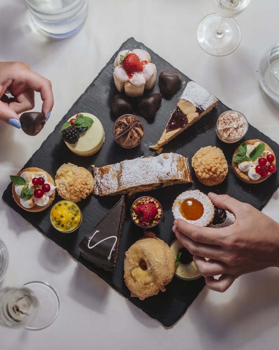 Vassoio nero con assortimento di dolcetti, mani che ne prendono alcuni, tavola bianca e bicchieri