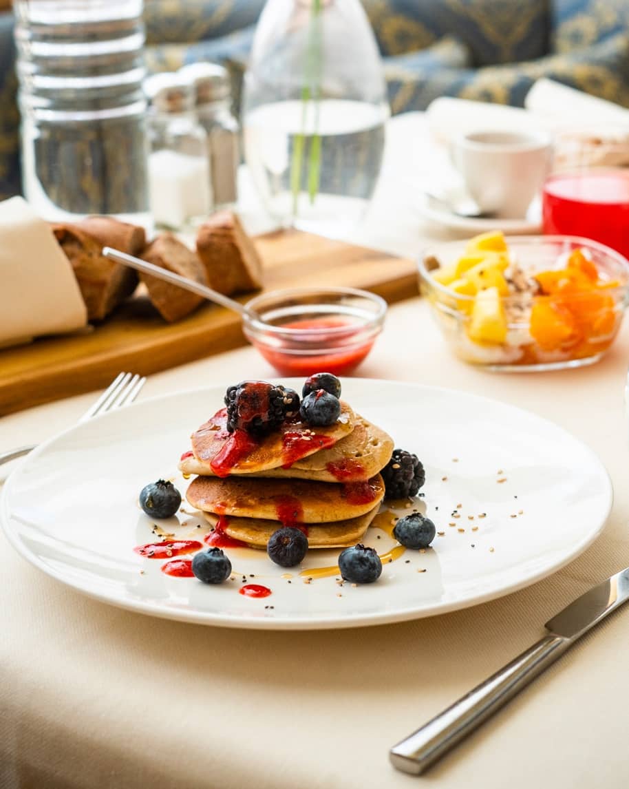 Tre pancake impilati con sciroppo e mirtilli su piatto bianco, tavola con pane, ciotola di frutta e posate