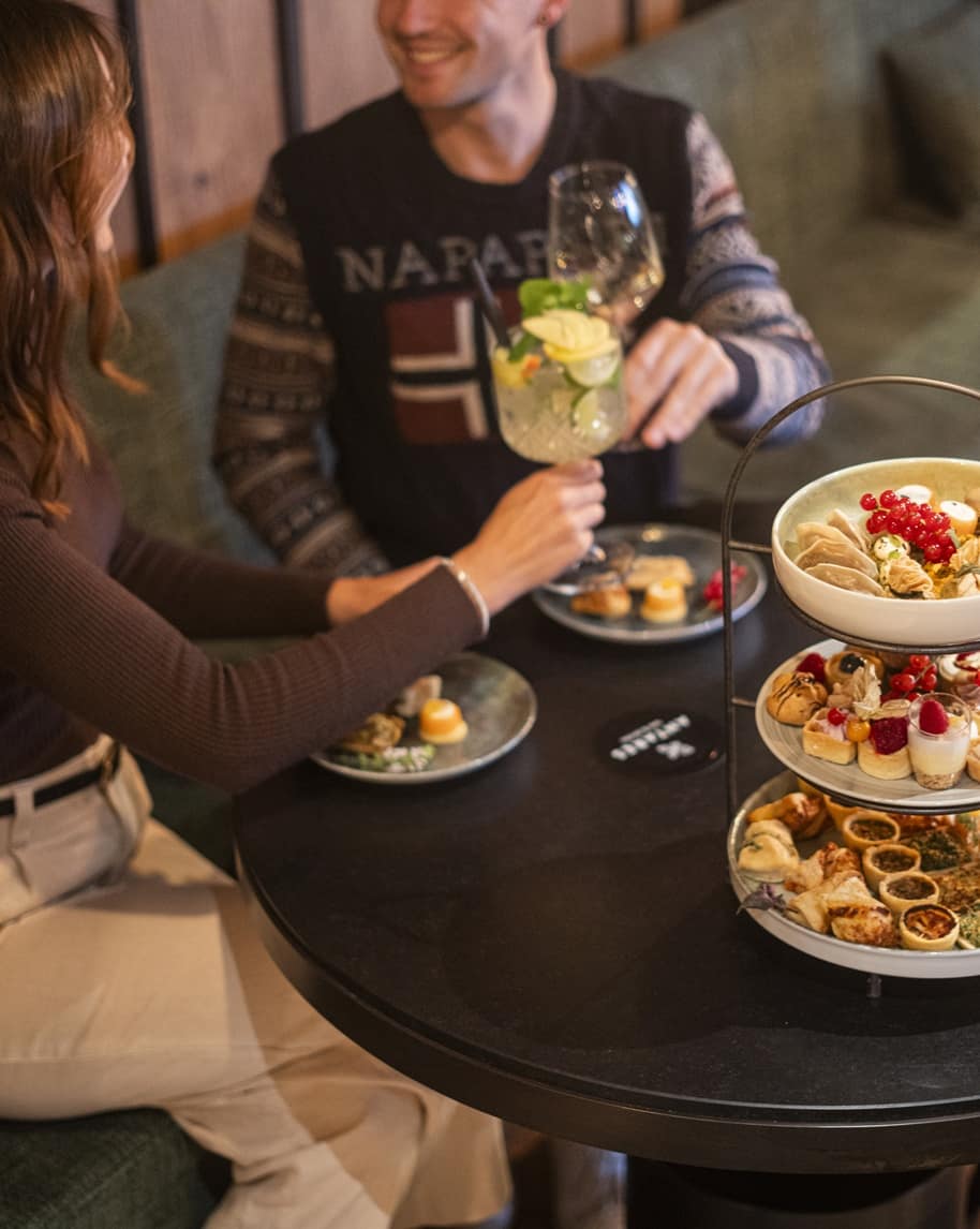 Due persone che brindano con bicchieri su tavolo con vassoio a tre piani di antipasti e dolcetti