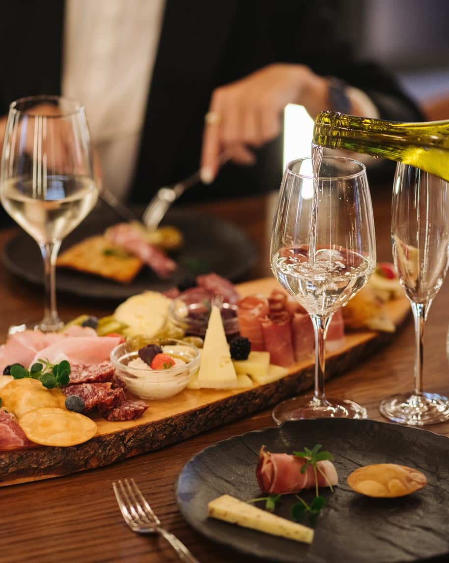 Tagliere di legno con salumi, formaggi e condimenti; calice mentre viene versato vino bianco, piatti sul tavolo