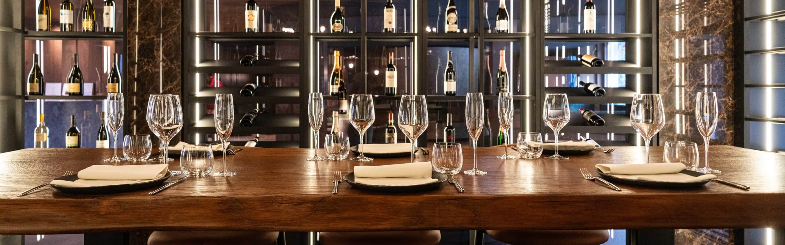 Tavolo di legno apparecchiato con piatti, bicchieri e posate davanti a scaffale con bottiglie di vino