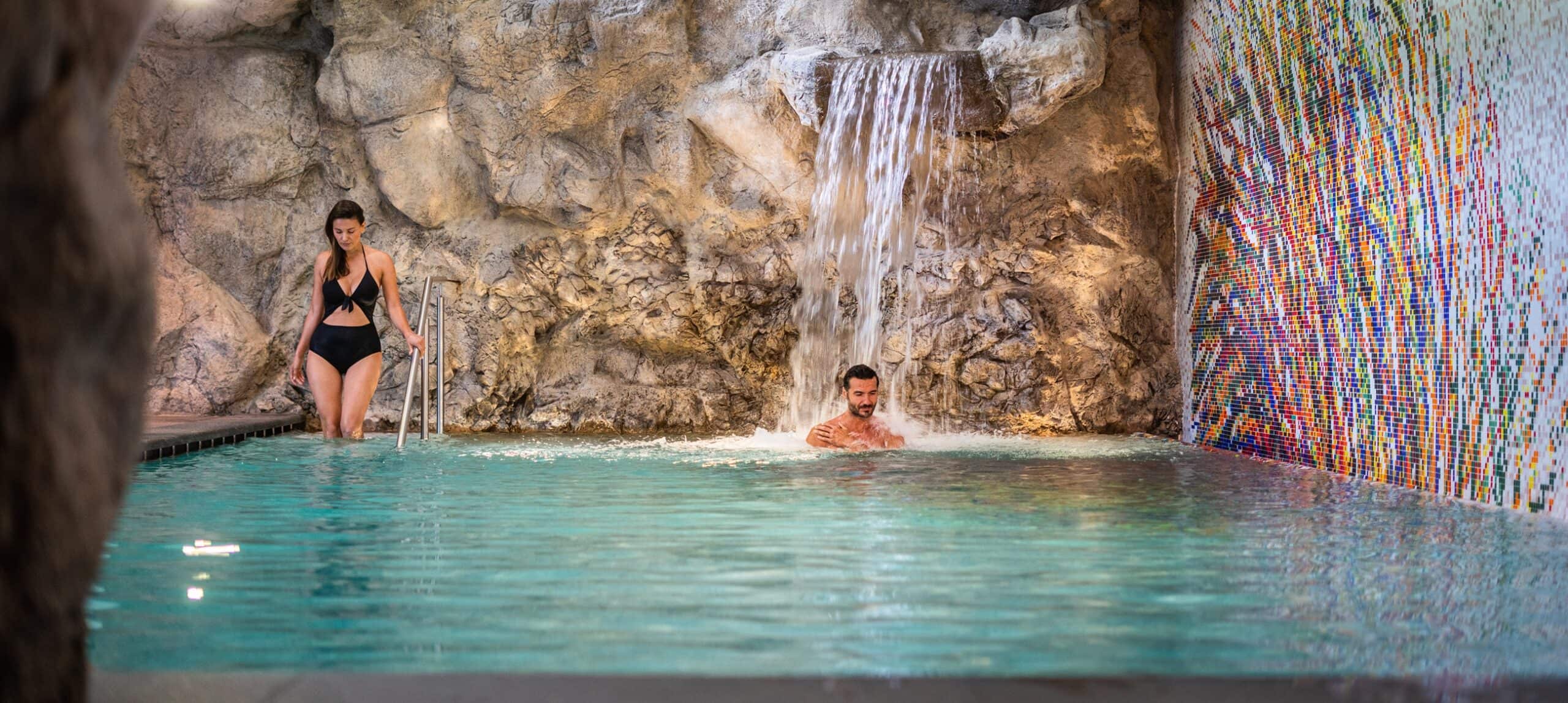 Donna scende in piscina dalla scaletta; uomo sotto cascata interna davanti a parete rocciosa e mosaico