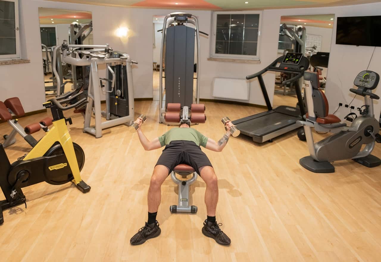 Uomo sdraiato su panca che solleva manubri a braccia aperte in palestra con attrezzi sullo sfondo