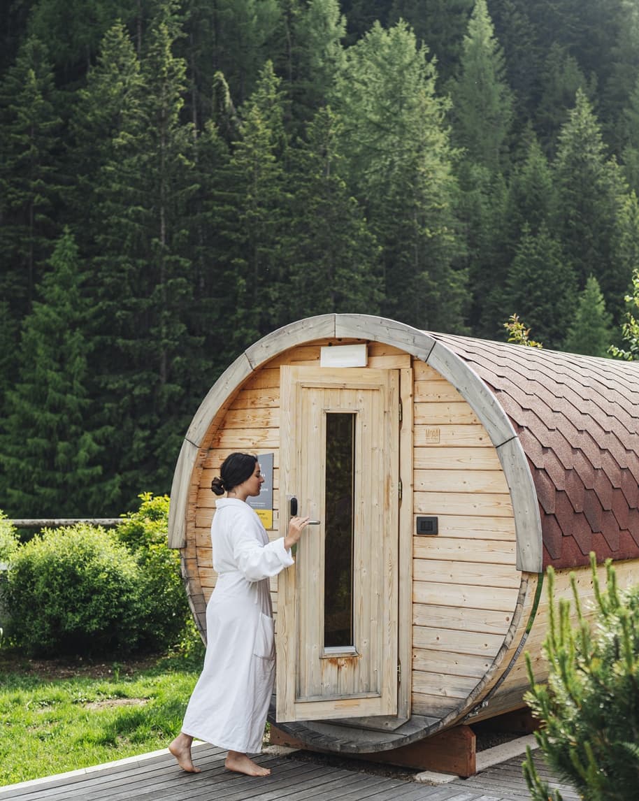 Persona in accappatoio entra in una sauna a botte in legno all'aperto, sullo sfondo foresta di conifere.