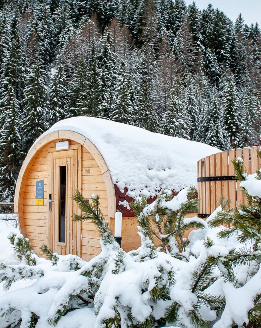 Cabina di legno a botte coperta di neve, rami innevati in primo piano e bosco sullo sfondo