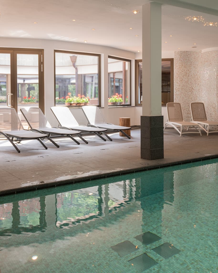 Piscina interna con lettini sdraio davanti a grandi finestre con vasi di fiori, pilastro centrale.