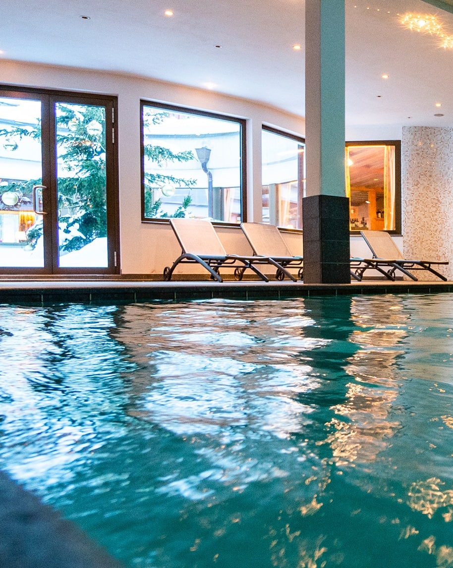 Piscina interna con superficie riflettente, lettini a bordo e ampie vetrate che scorgono alberi e neve