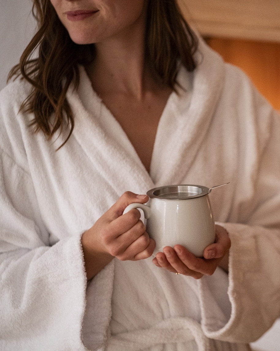 Persona in accappatoio bianco tiene una tazza bianca con infusore, mani in primo piano, ambiente interno