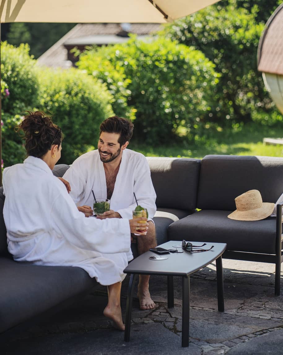 Uomo e donna in accappatoi su divano all'aperto che conversano sorseggiando bevande, tavolo con occhiali e cellulare