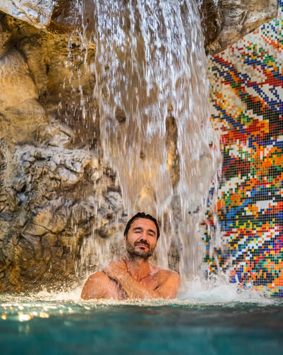 Uomo in piscina sotto cascata, parete rocciosa e mosaico a piastrelle colorate